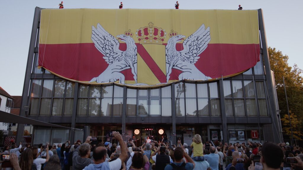 Höhenretter entrollen eine riesige badische Fahne mit Greifen und Wappen an der Fassade des Rathauses Achern, davor jubelndes Publikum