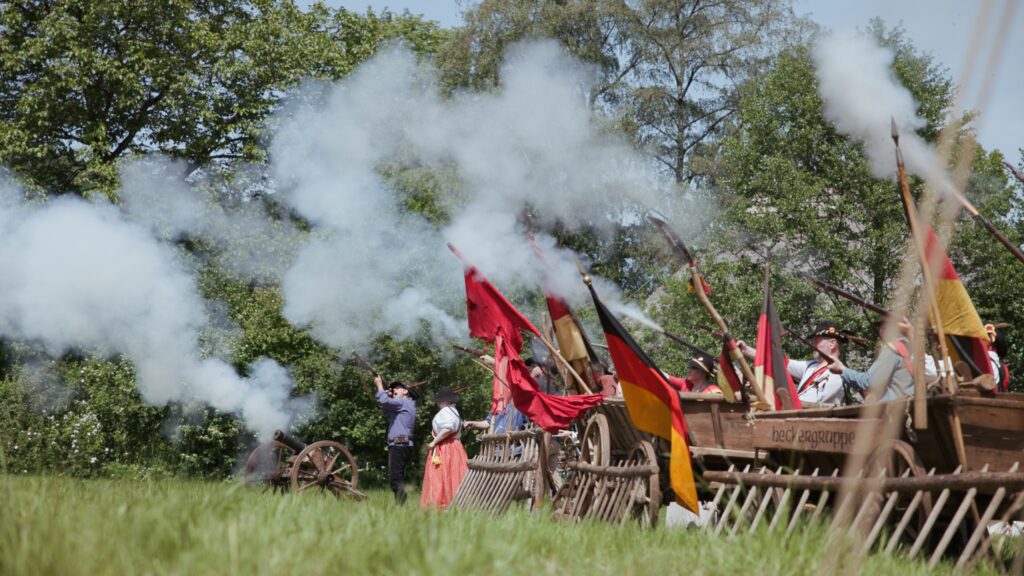Freischärler-Darsteller feuern hinter Leiterwagen eine Salve ab, Pulverdampf, schwarz-rot-goldene und rote Fahnen über der Wiese