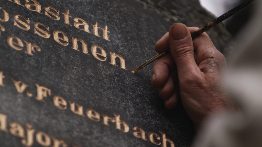 Hand zieht mit einem Pinsel die goldene Inschrift auf einem dunklen Gedenkstein für die in Rastatt Erschossenen nach