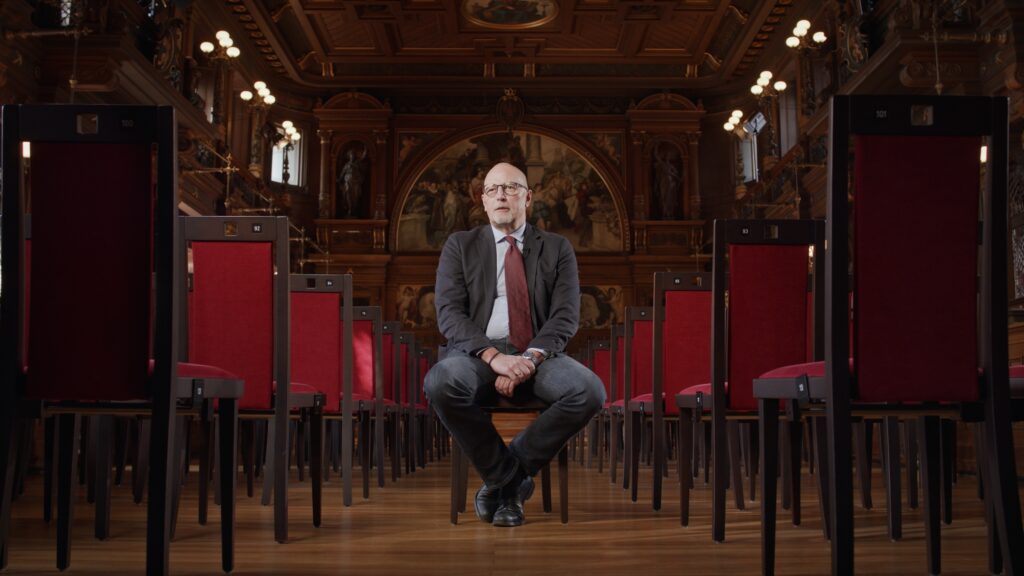 Interviewpartner sitzt zwischen Reihen roter Stühle in einem holzgetäfelten historischen Festsaal