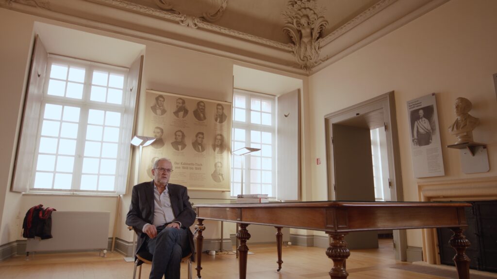 Interviewpartner sitzt in einem Ausstellungsraum neben einem Tisch, dahinter die Tafel „Der Kabinetts-Tisch von 1848 bis 1849“ mit Porträts