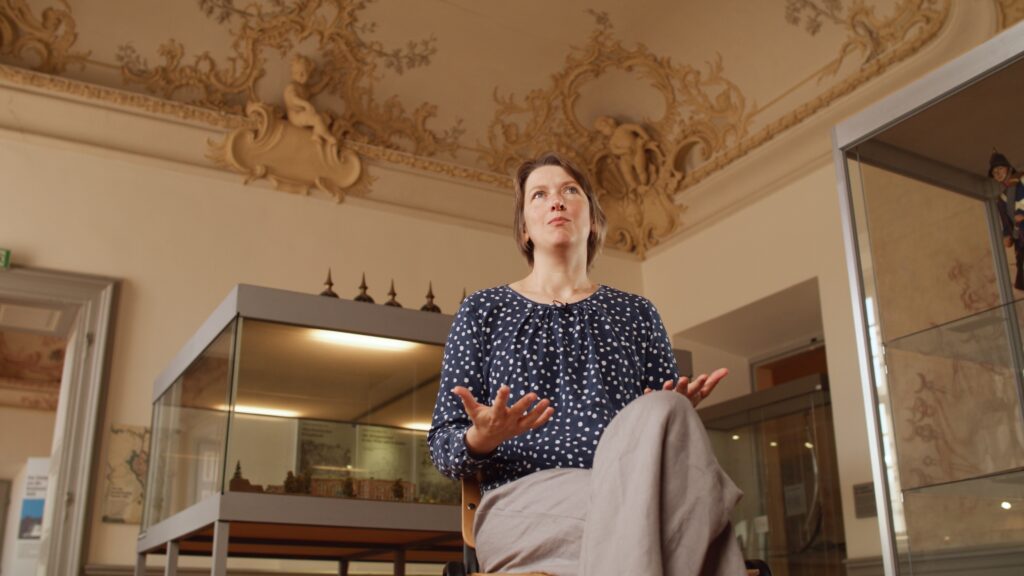 Interviewpartnerin spricht in einem Museumssaal mit Stuckdecke, hinter ihr Vitrinen mit einem Schlossmodell