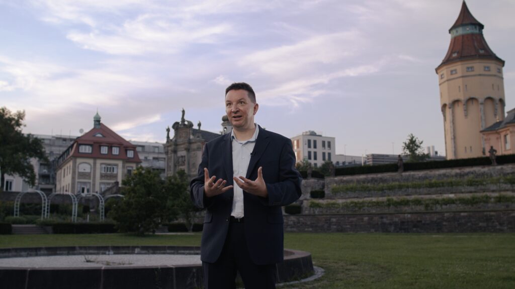 Interviewpartner spricht im Freien vor einem Terrassengarten und einem historischen Wasserturm