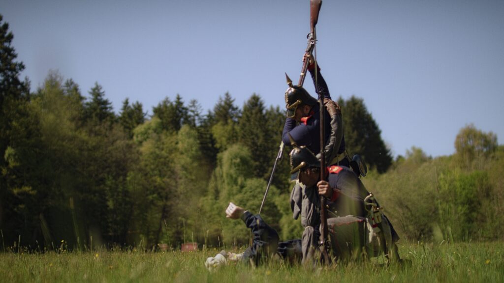 Zwei Soldaten mit Pickelhauben überwältigen auf einer Wiese einen am Boden liegenden Mann, einer holt mit dem Gewehrkolben aus
