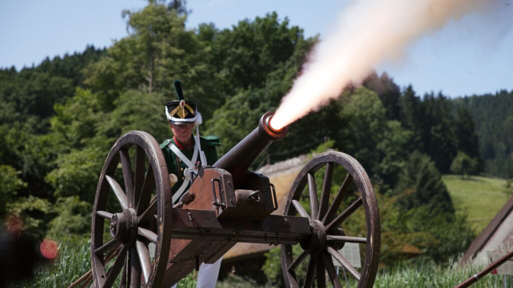 Kanonier in grüner Uniform mit Tschako hinter einer feuernden Feldkanone, Mündungsfeuer vor bewaldeten Hügeln