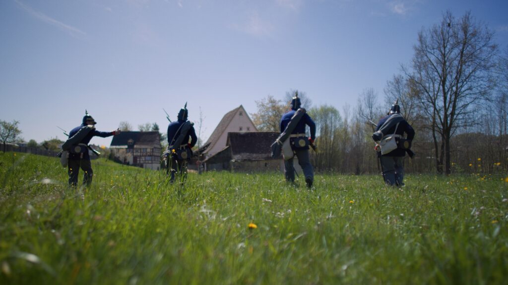 Vier Darsteller preußischer Soldaten mit Pickelhauben und Gewehren laufen über eine Frühlingswiese auf Fachwerkhäuser zu