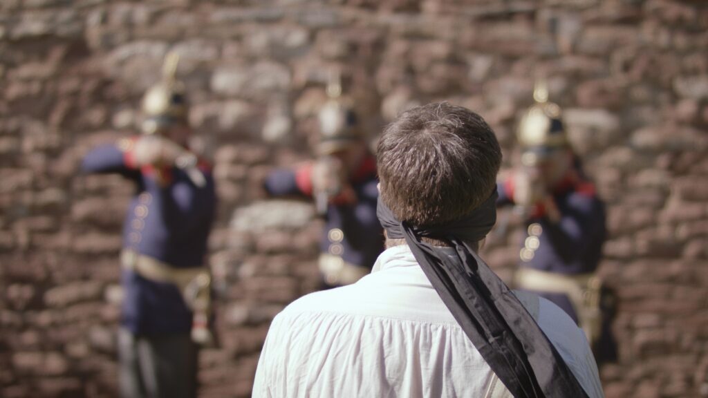 Mann mit Augenbinde steht vor einer Bruchsteinmauer, vor ihm legen drei Soldaten mit Pickelhauben die Gewehre an